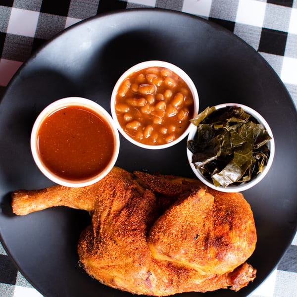 BBQ chicken with baked beans, collard greens, and sauce on a black plate.
