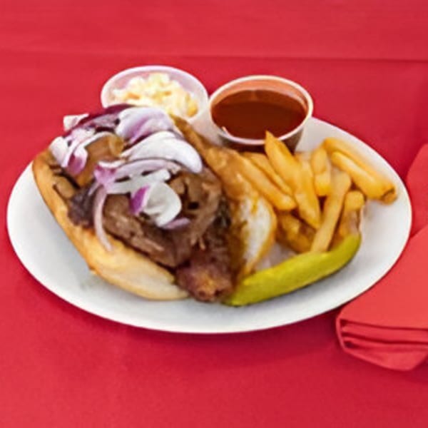 BBQ brisket sandwich with red onions, fries, coleslaw, and BBQ sauce on a white plate.